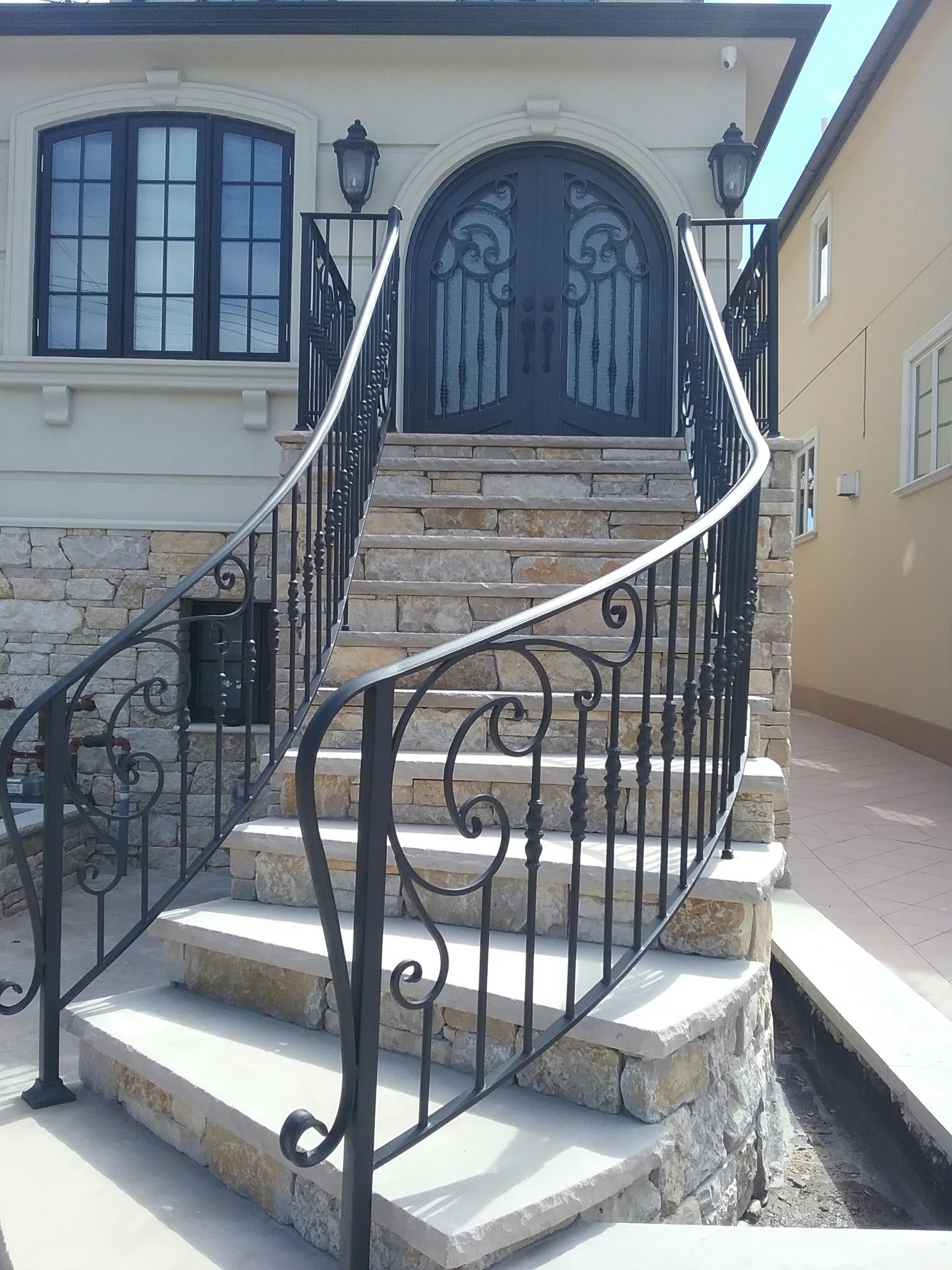 A grand curved exterior staircase with hand-forged scroll wrought iron railings leading up to an arched double iron door, a completed JBJ Iron Works installation